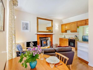 A living room with a fireplace, a dining table with a vase of flowers, a sofa, and a kitchen with wooden cabinets and a stove. - 1 Bed in Nethy Bridge (oc-28823) (Nethybridge)