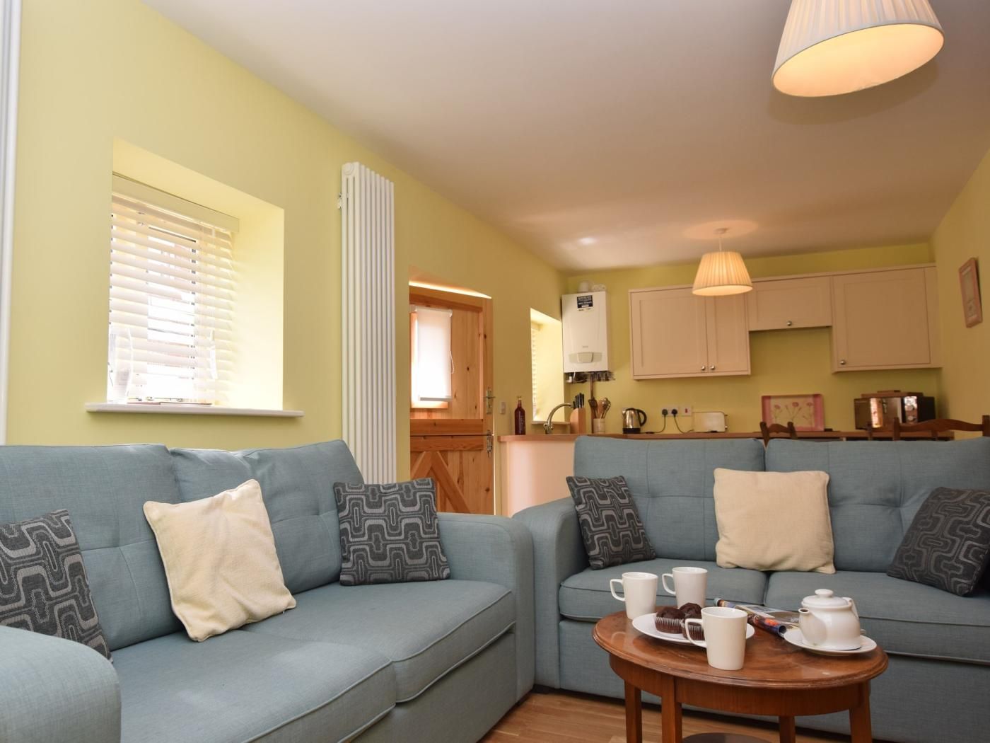A cozy living area with a sofa set, a small table with a teapot and cups, and a kitchen area in the background.