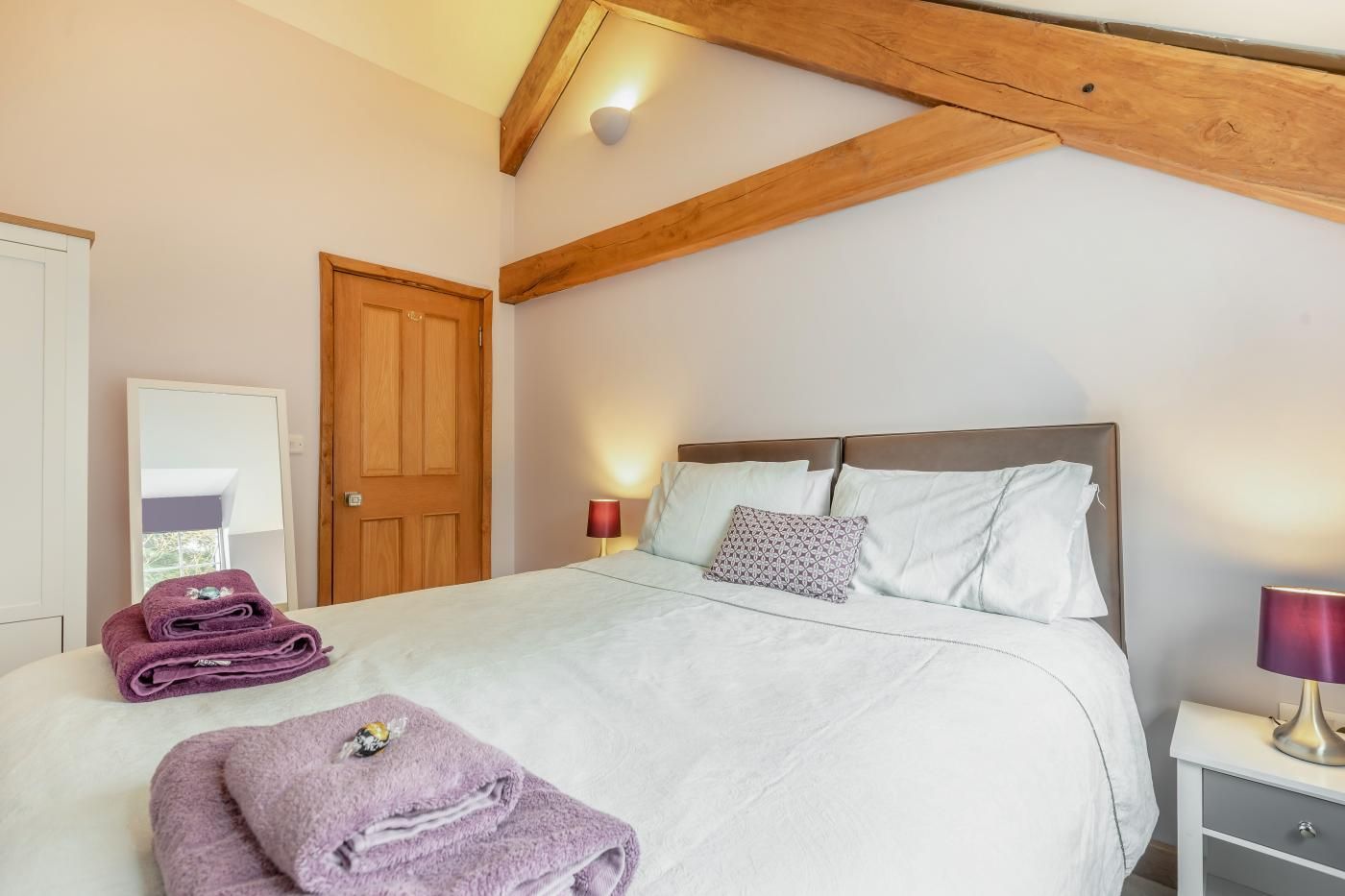 Een slaapkamer met een bed, kussens, een nachtkastje, een lamp, een deur en een raam.