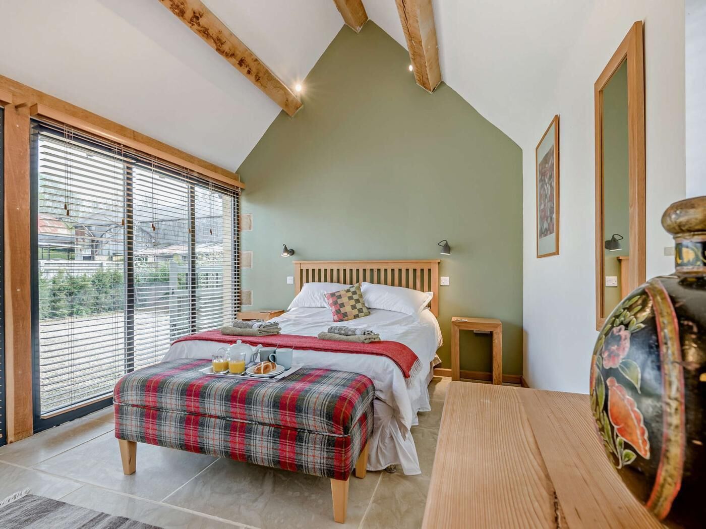 A bedroom with a bed, bedside tables, a bench, a window with blinds, and a view of the outside.