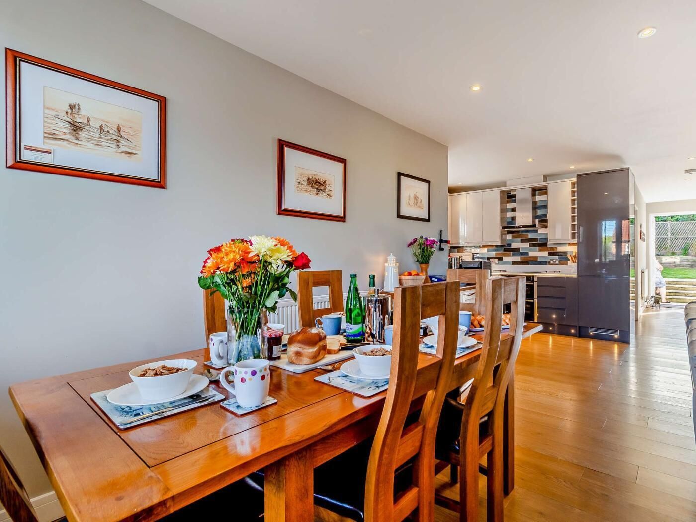 A dining area with a wooden table, chairs, and a vase of flowers. There are framed pictures on the wall and a kitchen in the background.