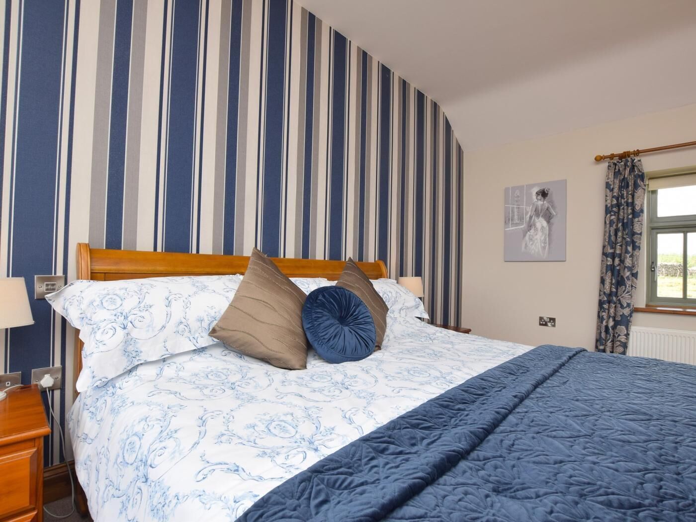 A hotel room with a striped wallpaper, a bed with patterned bedding, a wooden headboard, a bedside table with a lamp, and a window with curtains.