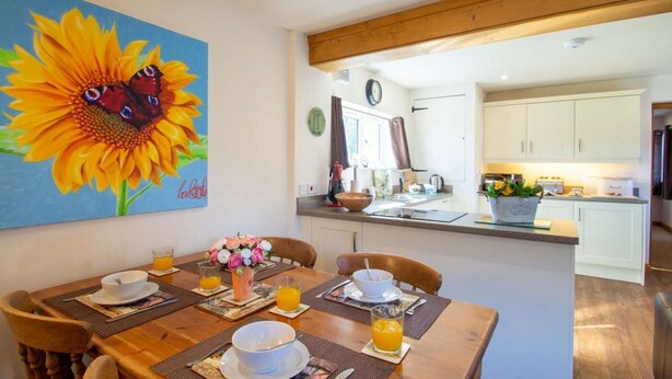 Un coin repas avec une table dressée pour quatre personnes, un tableau représentant un tournesol et, au fond, une cuisine.