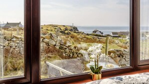 Vista a través de una ventana que muestra un muro de piedra, una colina cubierta de hierba y una masa de agua en la distancia.