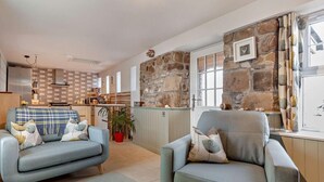 A modern living room with two grey armchairs, a sofa, a kitchen in the background, and a stone wall. - 1 Bed in Forres (oc-32129) (Alves)