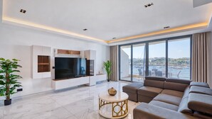 A modern living room with a flat-screen TV, a sectional sofa, a coffee table, and a large window offering a city view. - Sliema 3BDR Flat with Seaview (Tas-Sliema)
