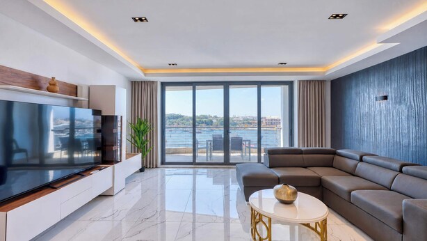 A modern living room with a large flat-screen TV, a sectional sofa, a round coffee table, and a view of the sea through large windows. - Sliema 3BDR Flat with Seaview (Tas-Sliema)