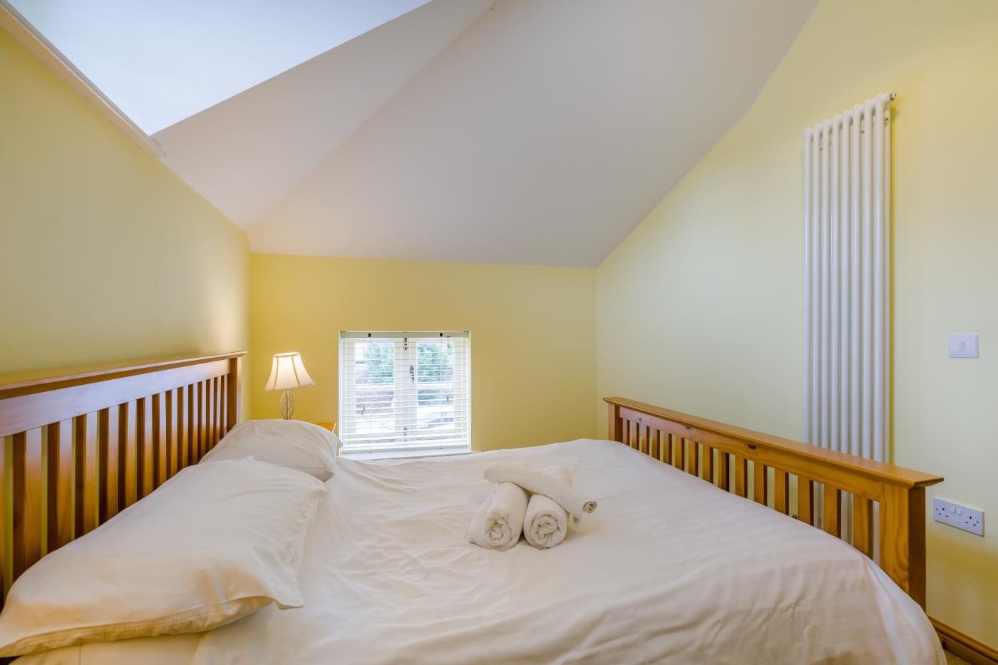 Une chambre avec un plafond en pente, un lit avec du linge de lit blanc, une fenêtre avec des stores et un radiateur.