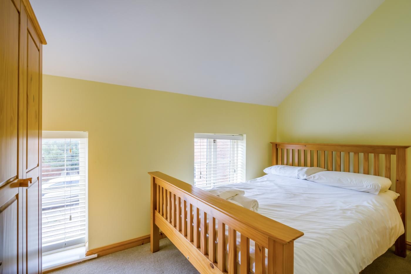 A bedroom with a wooden bed, two windows with blinds, and a wooden wardrobe.