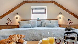 Een slaapkamer met een bed, kussens, een houten hoofdbord, een raam, wandlampen, een decoratief paardbeeldje, en een dienblad met croissants, sap en koffiekopjes.