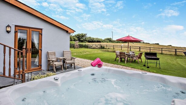 Een bubbelbad met houten stoelen en een rode parasol, een houten terras en een grasveld op de achtergrond.