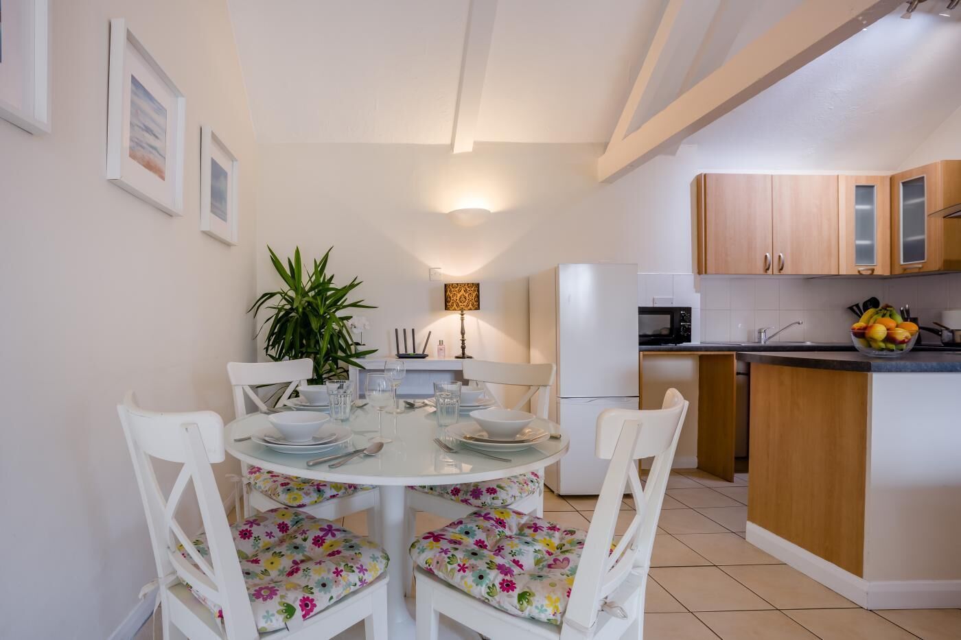 A dining area with a round table, white chairs with floral cushions, a potted plant, and a wall-mounted light fixture.