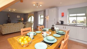 Uma sala de jantar com mesa de madeira posta para quatro, cozinha com armários brancos e sala de estar com sofá e cortinas.