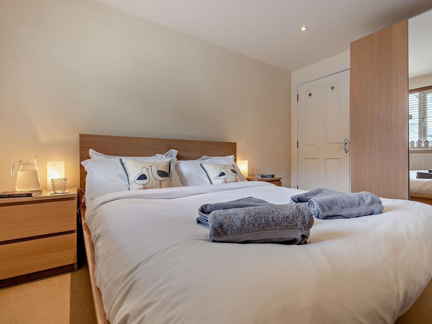 A neatly made bed with pillows, a bedside table with a lamp, and a nightstand with a water pitcher.