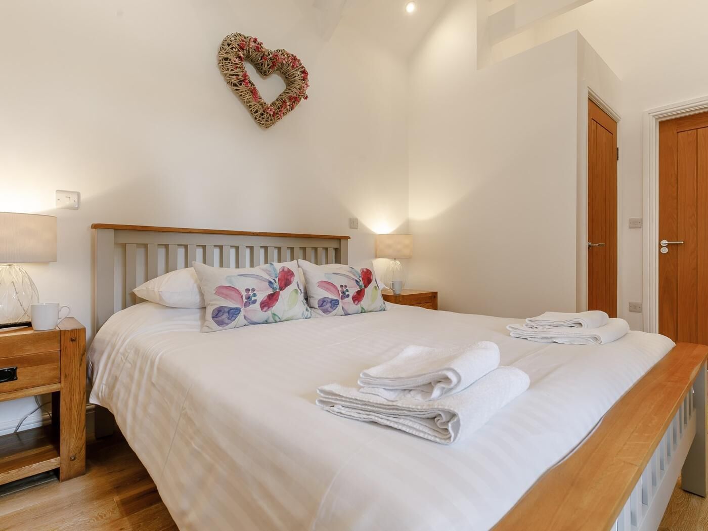 A bedroom with a bed, pillows, a heart-shaped decoration, a nightstand, a lamp, and a door.