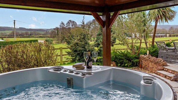 Ett bubbelbad med utsikt över ett lantligt landskap, en träpergola och utemöbler.