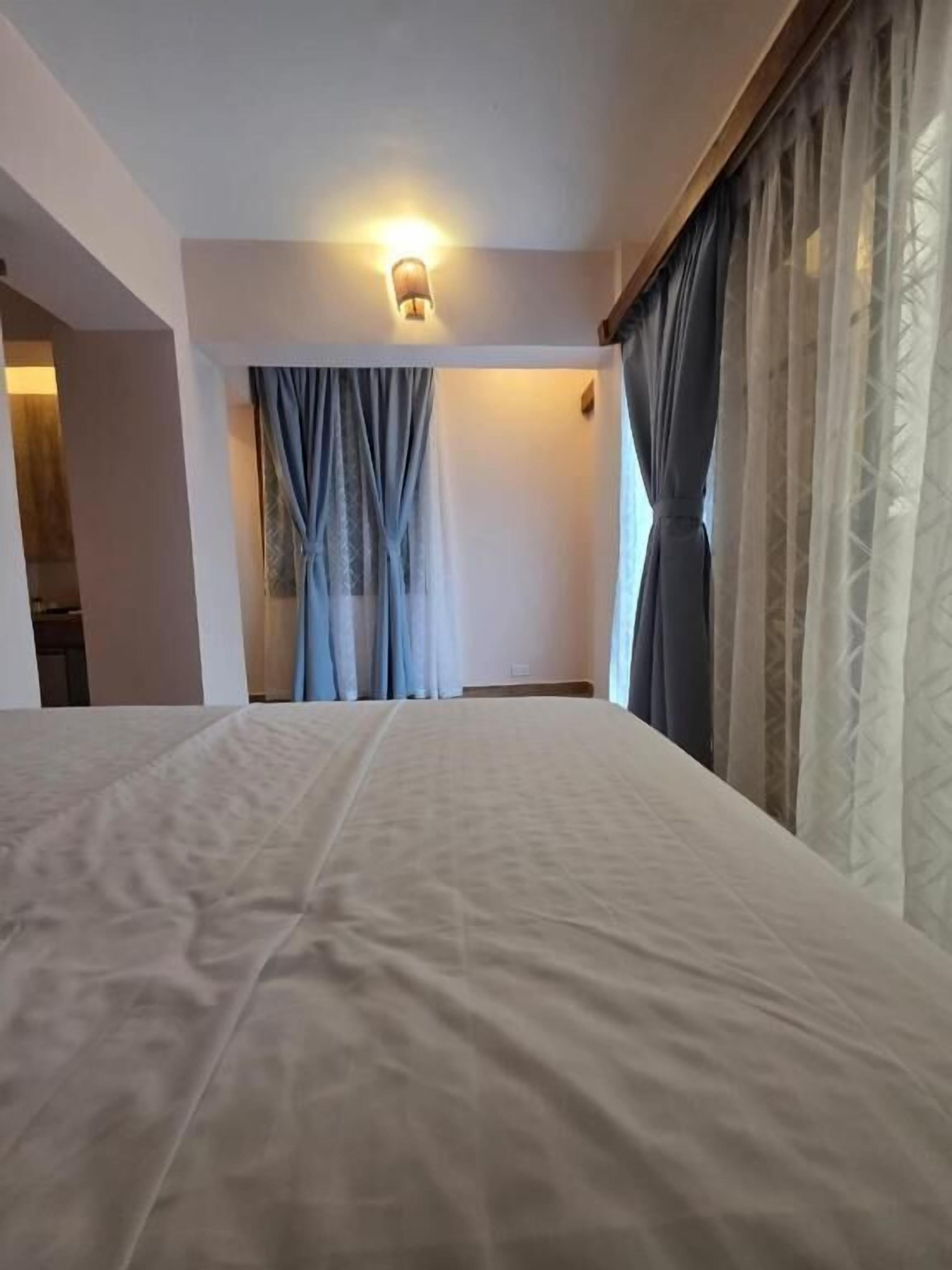Una habitación de hotel con cama, cortinas, una lámpara fijada a la pared y un ventanal con cortinas.