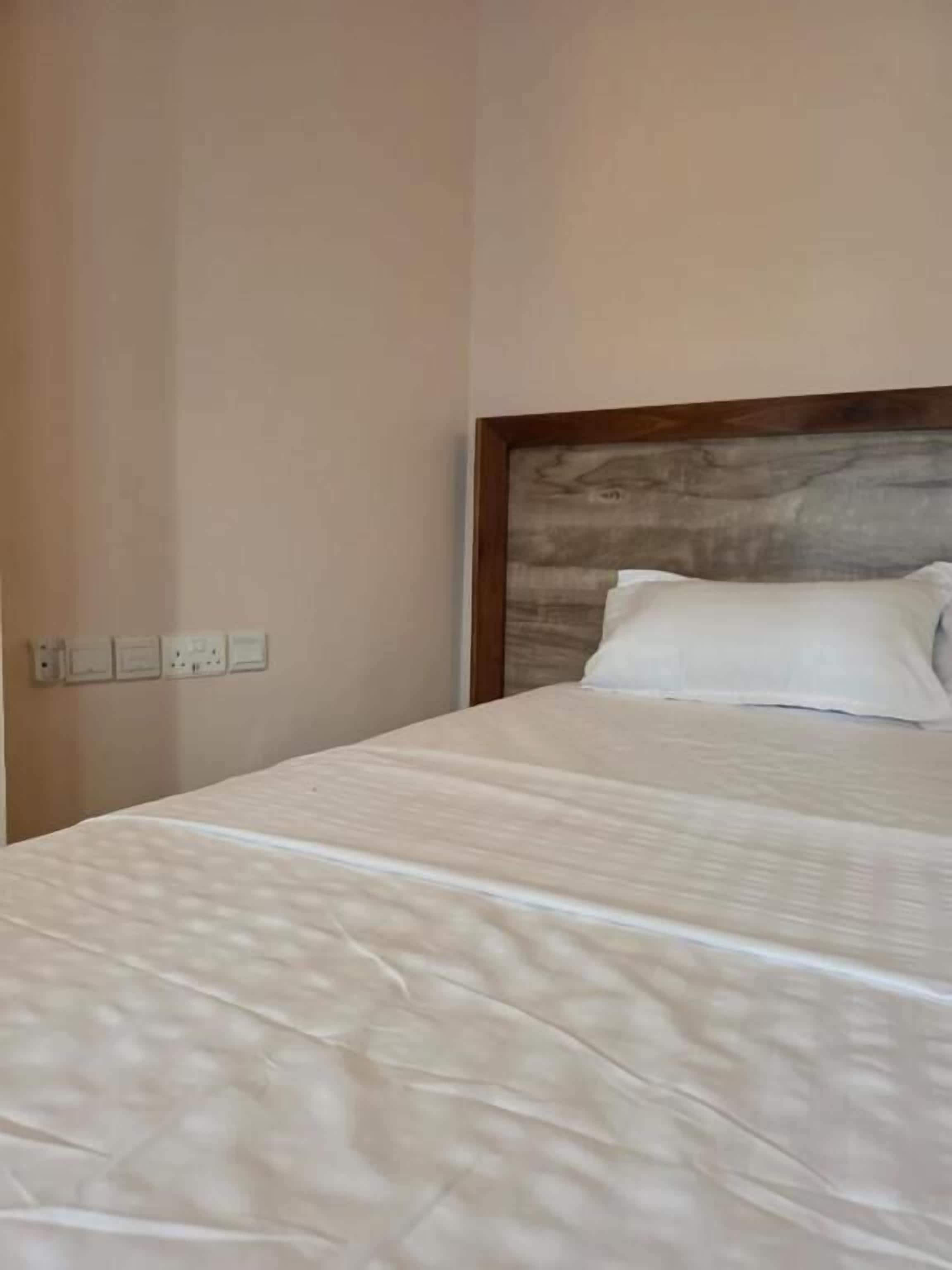 A bed with a wooden headboard, a white pillow, and a plain wall with a power outlet.