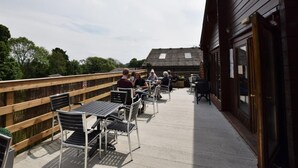 Eine Terrasse im Freien mit Metalltischen und -stühlen, einem Holzzaun und einem Gebäude mit geneigtem Dach.