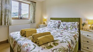 A bedroom with a floral bedspread, two rolled towels on the bed, a nightstand with a lamp, and a window with curtains. - 2 Bed in Bideford (oc-36456) (Weare Giffard)