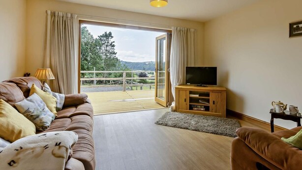 A living room with a sofa, television, and a view through a glass door to an outdoor deck. - 2 Bed in Bideford (oc-36456) (Weare Giffard)