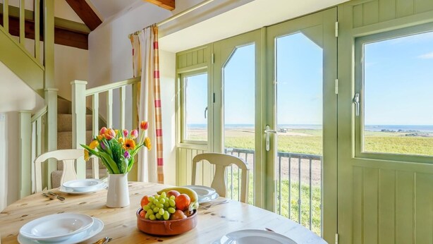 Un coin repas avec une table dressée pour quatre personnes, un vase de fleurs et une vue sur la plage par de grandes portes ouvertes.