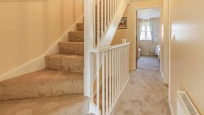 Un escalier recouvert de moquette, avec des rampes blanches, mène à une chambre dotée d’une fenêtre et d’un radiateur.