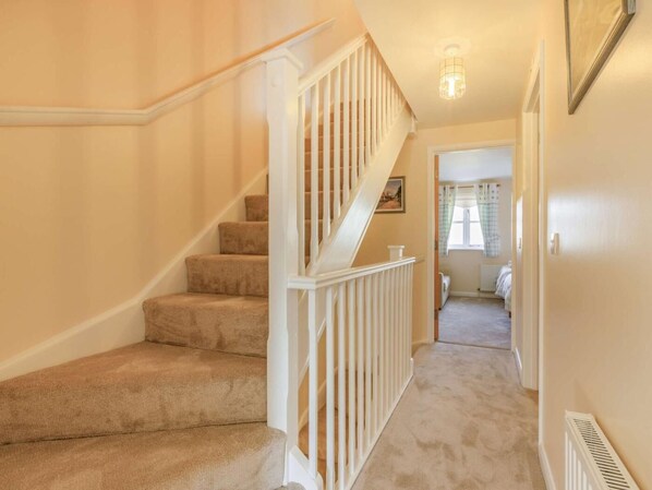 Un escalier recouvert de moquette, avec des rampes blanches, mène à une chambre dotée d’une fenêtre et d’un radiateur.