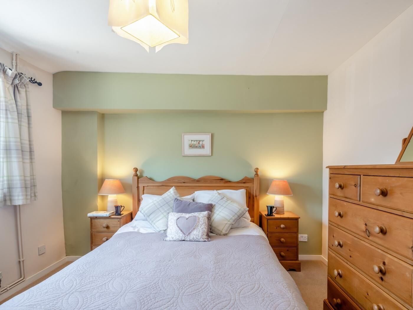 A bedroom with a bed, two bedside tables, a dresser, a wall-mounted lamp, and a framed picture.