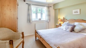 Une chambre à coucher avec un lit en bois, une chaise en osier, une fenêtre avec des rideaux, un radiateur et une table de chevet avec une lampe.