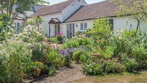 Un bâtiment blanc, doté d’un toit en tuiles, entouré d’un jardin où poussent différentes plantes et fleurs.