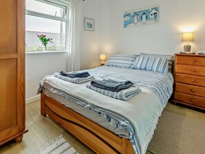 A neatly made bed with striped bedding, a wooden dresser, a bedside lamp, and a window with a view of a building and flowers. - 2 Bed in Helston (oc-36430) (Mullion)