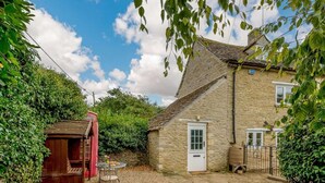 Une maison en pierre avec une porte blanche, un petit jardin avec un banc en bois et une cabine téléphonique rouge.