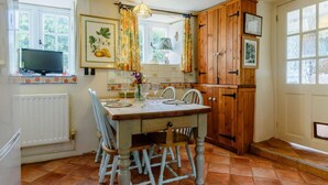 Une cuisine avec une table à manger, des chaises, des meubles de rangement en bois, une fenêtre avec des rideaux et un radiateur.