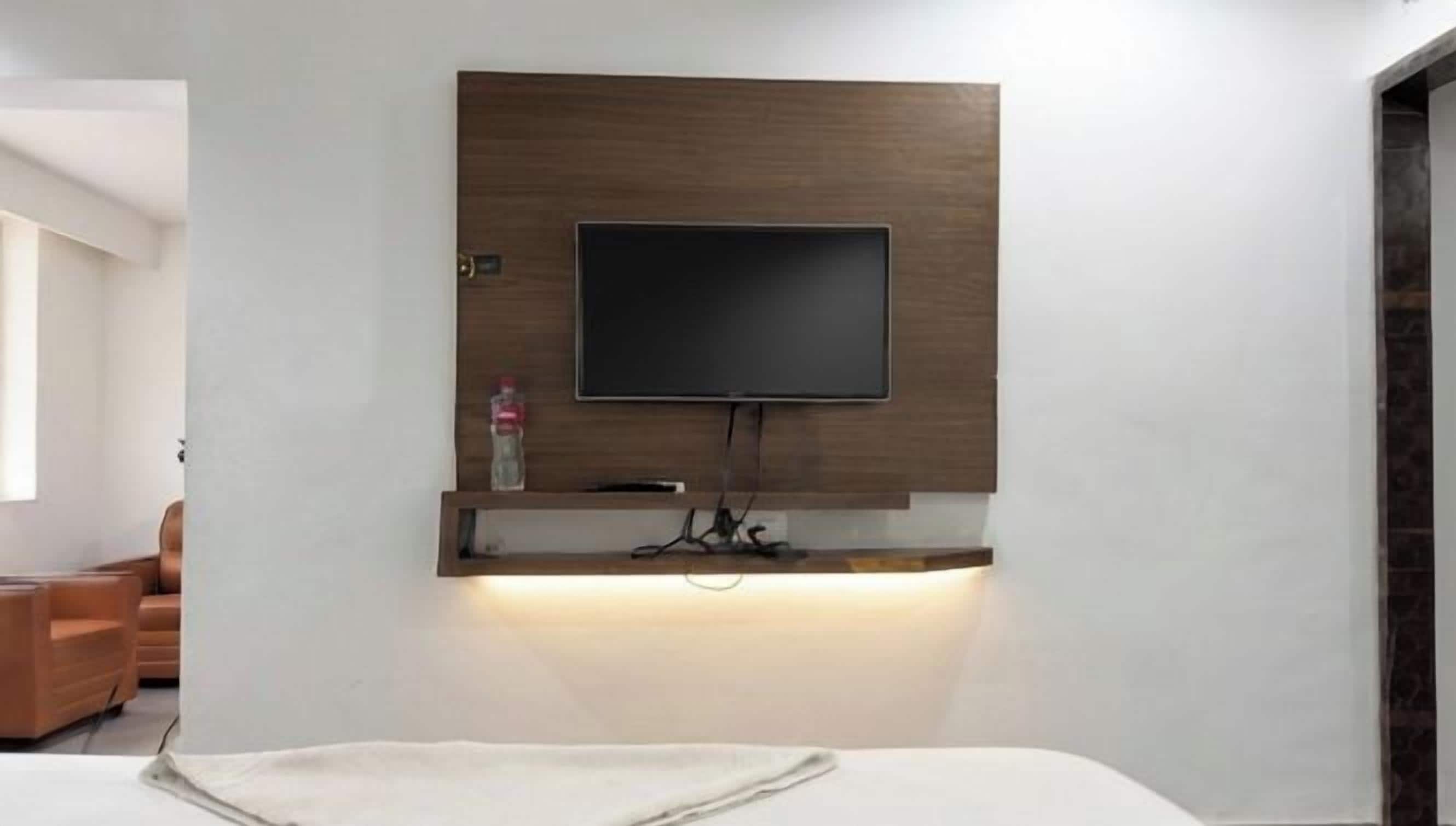 Habitación de hotel moderna con una televisión de pantalla plana montada, un estante de madera debajo y una cama con una colcha blanca.