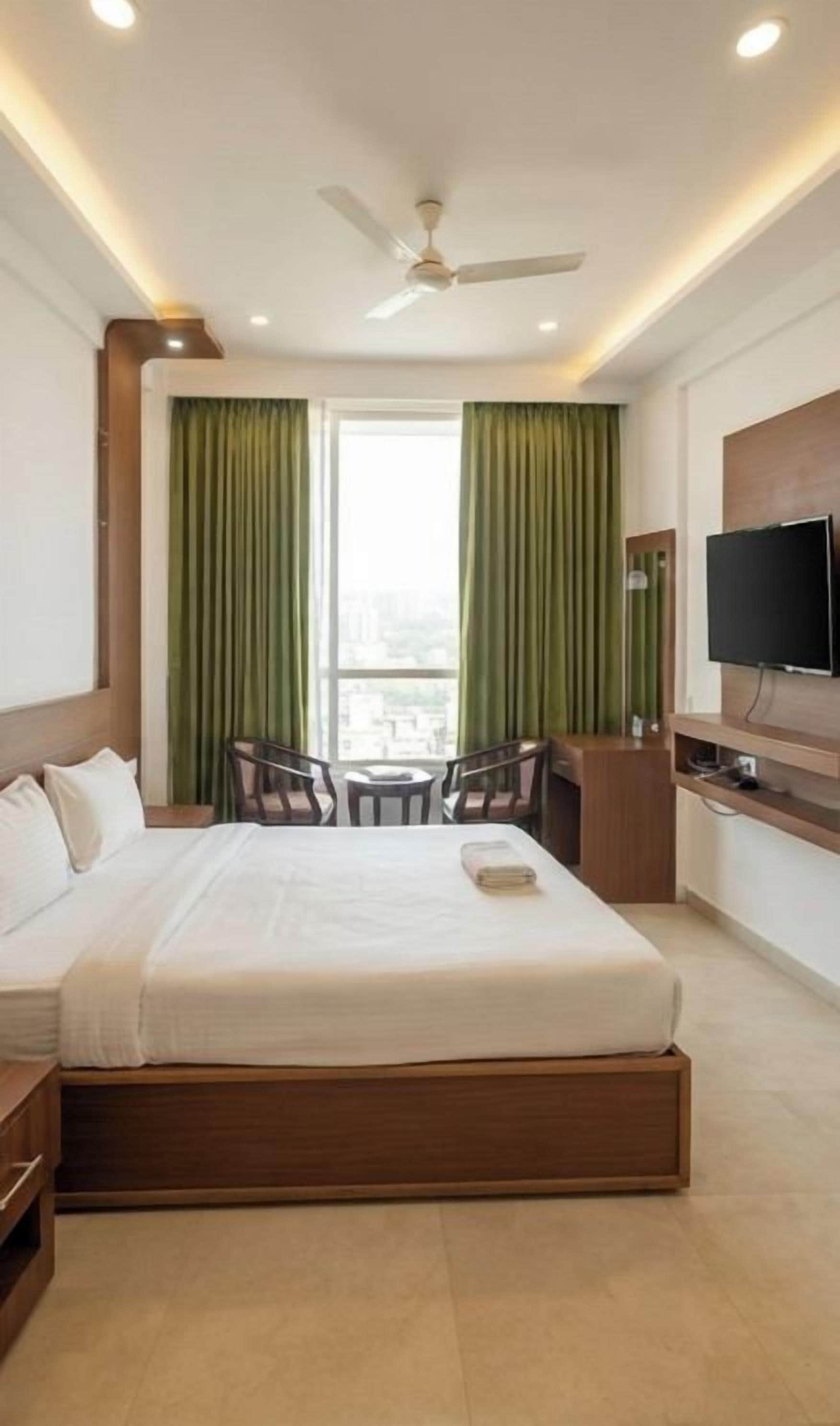 Habitación de hotel con cama, escritorio, silla, televisión y ventana con cortinas.