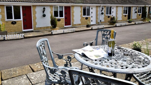 Uma fileira de casas tradicionais com portas vermelhas e janelas brancas, uma rua pavimentada e uma mesa de metal com cadeiras em primeiro plano.