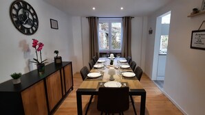 A dining area with a wooden table set for four, a black chair, a clock, and a window with curtains. - Large, renovated house in central Alsace (130 m²) (Neubois)
