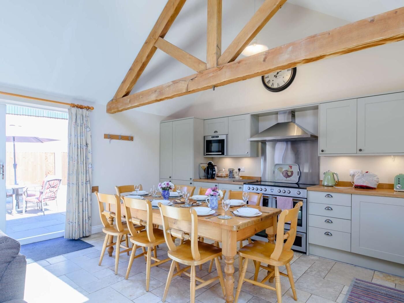 A dining area with a wooden table and chairs, a kitchen with white cabinets and appliances, and a clock on the wall.