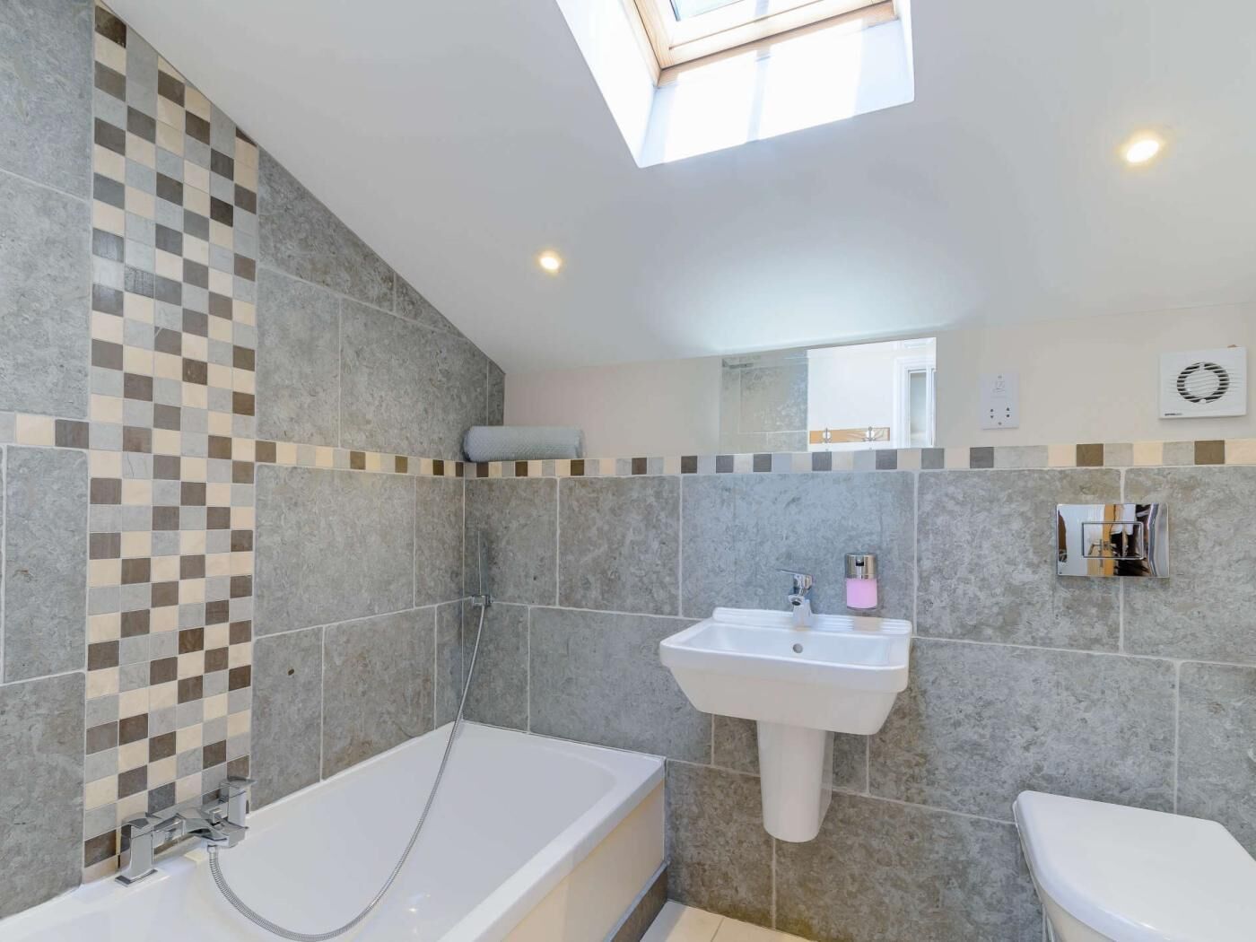 A modern bathroom with a bathtub, sink, and toilet, featuring grey tiles and a skylight.