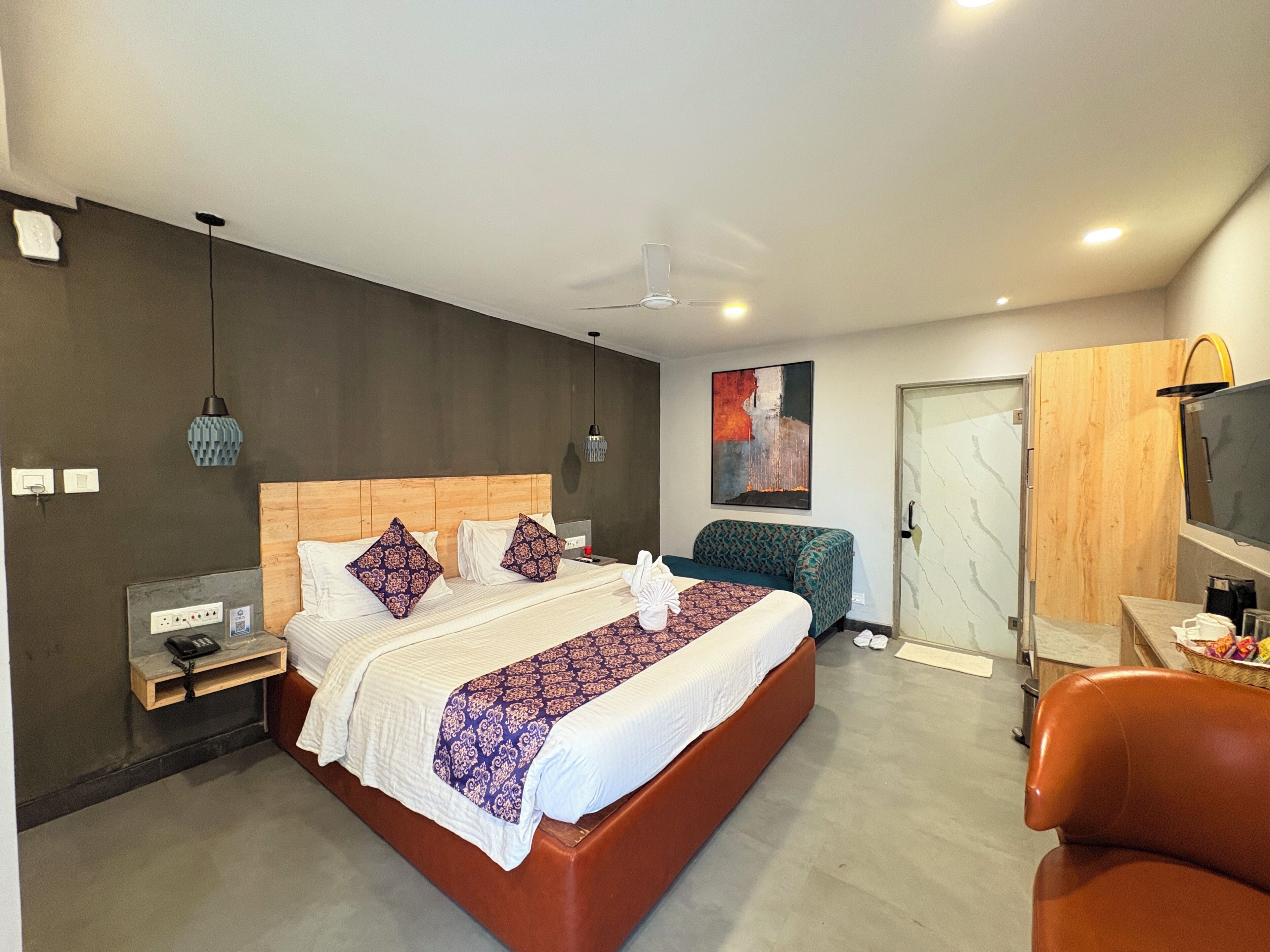 A modern hotel room with a large bed, a wooden headboard, a television, and a seating area with a leather chair.