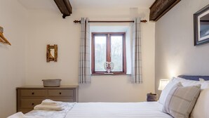 Un dormitorio con cama, una ventana con cortinas, una mesita de noche con cesta y un cuadro colgado en la pared.