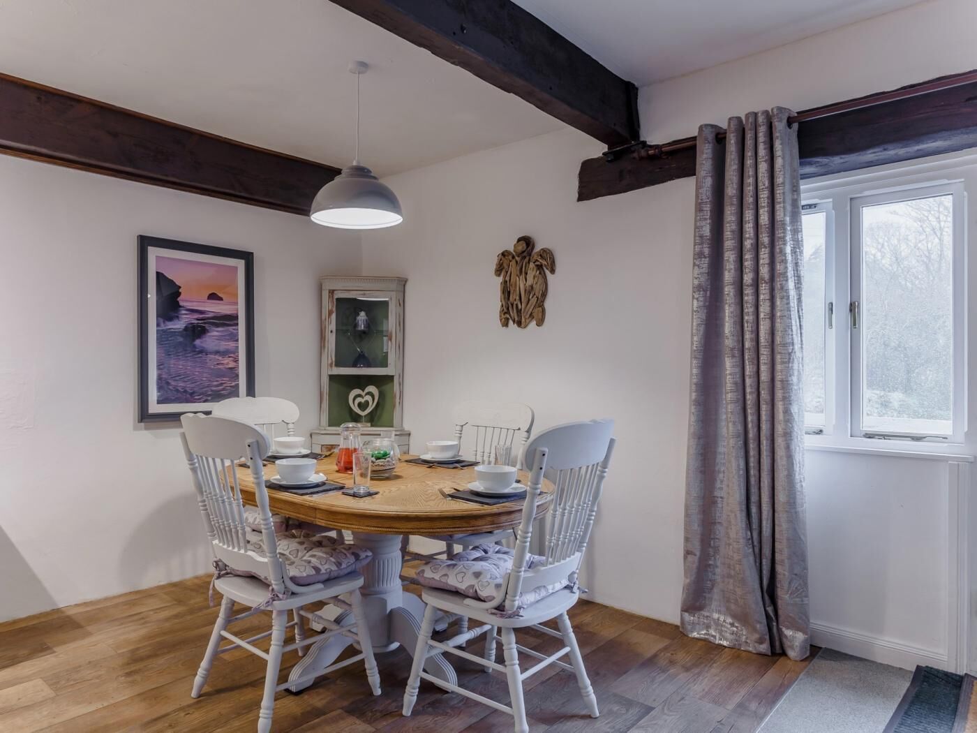 Uma sala de jantar com uma mesa redonda, cadeiras brancas, uma luminária pendente, um quadro emoldurado e uma janela com cortinas.
