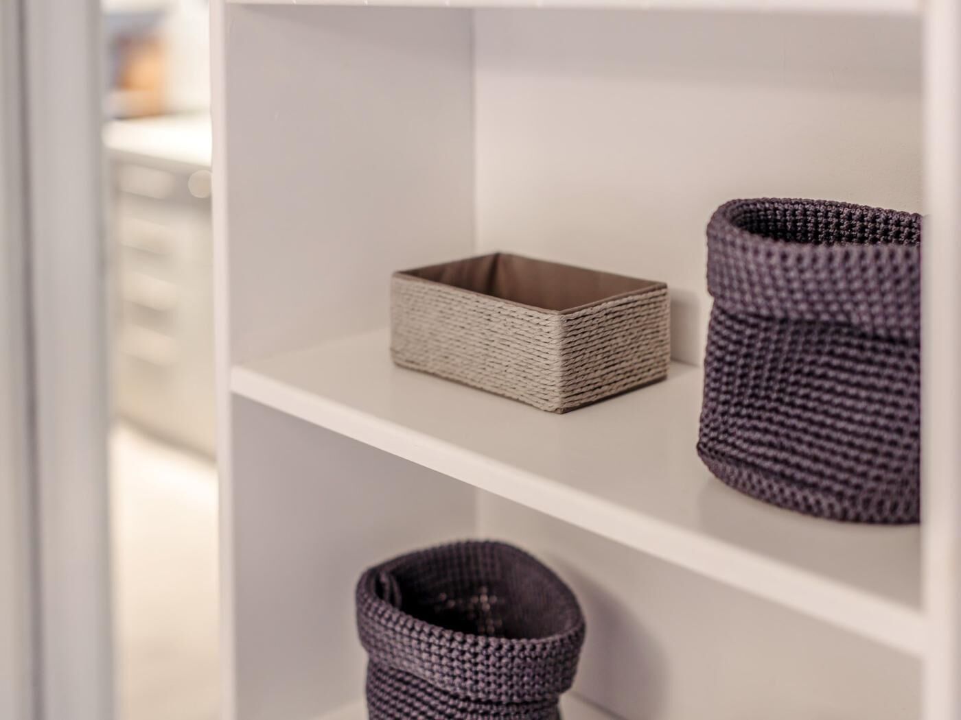 Een wit schap met drie gevlochten manden in verschillende kleuren en maten.