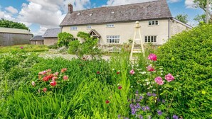 A stone house with a sloped roof, surrounded by a garden with various flowers and a wooden ladder.