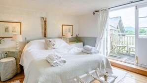 A bedroom with a large bed, two bedside tables with lamps, a window with a view of a wooden deck and mountains, and a chair by the window.
