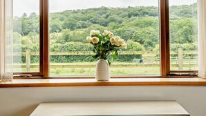 Une fenêtre donnant sur un paysage verdoyant, avec un vase blanc rempli de fleurs posé sur le rebord de la fenêtre.
