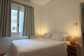 Une chambre à coucher avec un lit, des rideaux, une table de chevet, une lampe et une fenêtre.