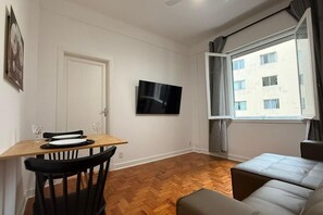 Une pièce avec un plancher en bois, une table à manger avec des chaises, un canapé, un téléviseur fixé au mur et une fenêtre avec des rideaux.
