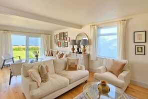 A spacious living room with a large sofa, a smaller armchair, a coffee table, and a dining area with a chair and table. - The Coast House (Porthcothan Bay)